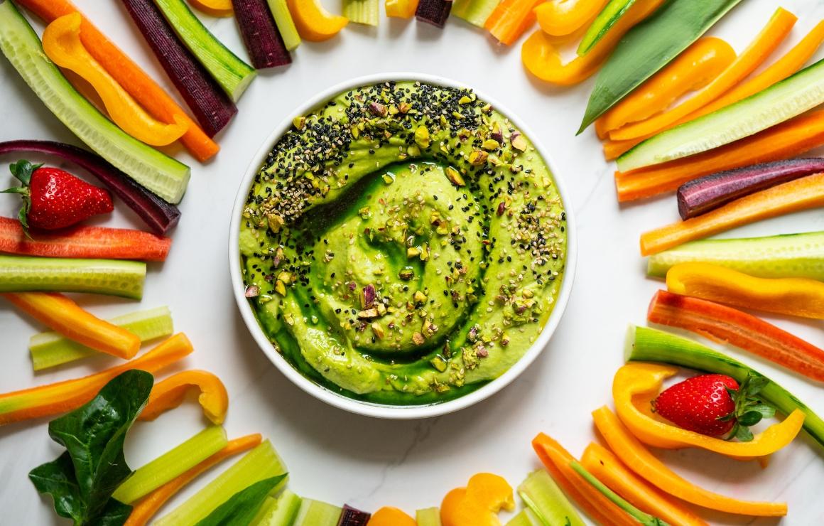 Bärlauch-Hummus in einer Schale mit Pistazien, hellem und schwarzem Sesam sowie einem Schuss grünem Bärlauch-Öl getoppt; drumherum drappiert Gemüsesticks aus Karotten, Sellerie, Paprika und Gurke