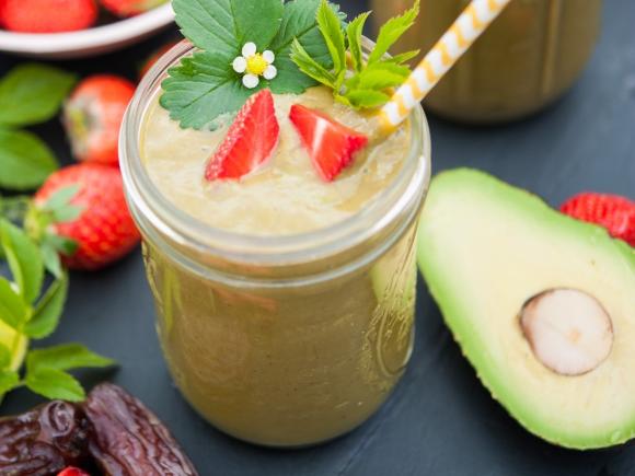 Grüner Smoothie, daneben eine aufgeschnittene Avocado sowie andere Früchte und Blattgrün.