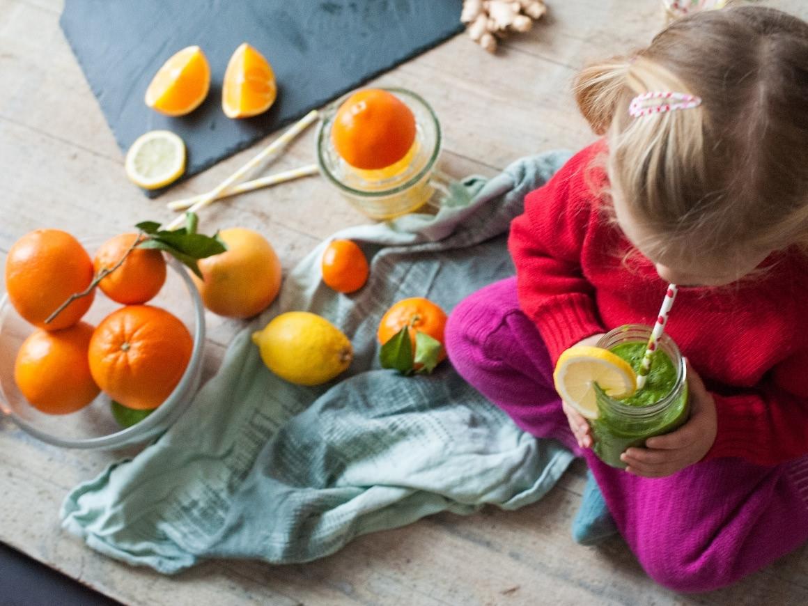 kleines Mädchen sitzt auf einem Tisch, trinkt einen Grünen Smoothie und Zitronenscheibe dekoriert. Auf dem Tisch liegen Orangen, Mandarinen, Zitronen.